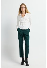 Camicia bianca a maniche lunghe con bottoni, abbinata a pantaloni scuri verdi su misura e stivaletti neri con tacco alto. Tessuto morbido, design aderente.