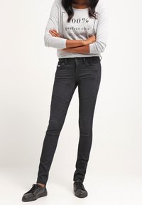 Jeans ajustados negros con detalles acolchados en las rodillas, combinados con una camiseta de manga larga gris claro que presenta texto negro y zapatillas negras.