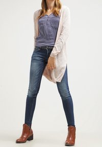 Světle růžový kardigan přes modrou košili, spárovaný s tmavými denimovými džíny a hnědými kotníkovými botami. Kardigan má dlouhé rukávy a otevřený přední díl.