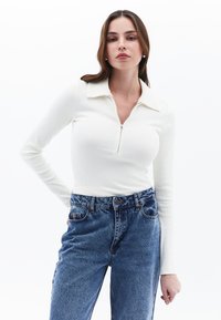 Biała bluzka z prążkowanej tkaniny z zamkiem przy kołnierzu, w połączeniu z niebieskimi dżinsami o wysokim stanie. Stylizacja ma swobodny krój i klasyczny design.