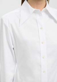 Camicia bianca con colletto ampio, caratterizzata da bottoni dorati e una texture liscia. Vista ravvicinata del colletto e dei bottoni superiori.