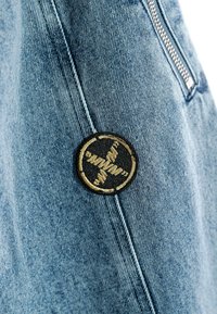 Tejido de denim en azul claro con costuras visibles, que presenta un parche bordado circular en negro y dorado en el lado. Detalle de cremallera presente.