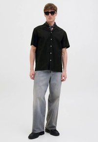 Melna īsroku krekla ar teksturētu rakstu, sapārota ar gaiši pelēniem platu kāju džinsiem un melnām masīvām kurpēm. Aksesuāri ietver melnus saulesbrilles.