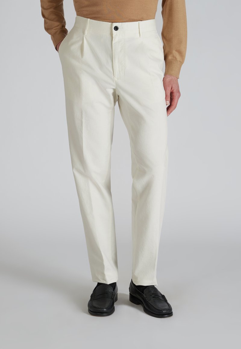 Pantalons blancs plissés en tissu de coton lisse, dotés d'une fermeture à un bouton et de poches latérales. Associés à des mocassins noirs.
