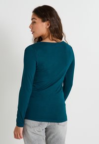 Camiseta de manga larga color verde azulado con cuello redondo, textura suave y diseño ajustado. Usada con pantalones gris claro, mostrando detalles sutiles de costura.