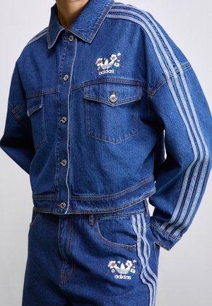 Denim Adidas Jacke und Hose mit drei hellen Streifen und Blumenlogos auf Brust und Oberschenkel.