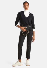 Kobieta w białym golfie, czarnym swetrze w serek, czarnych spodniach, białych trampkach, trzymająca czarną torebkę z złotym łańcuchem na talii.