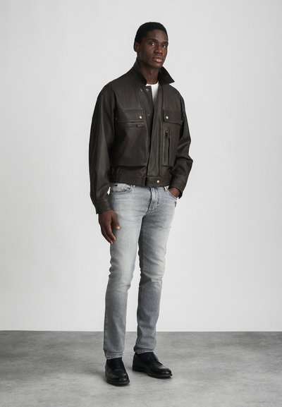 Καφέ δερμάτινο μπουφάν τύπου bomber με τέσσερις μπροστινές τσέπες και χρυσά αξεσουάρ, φορεμένο πάνω από ένα λευκό πουκάμισο, συνδυασμένο με ανοιχτό γκρι τζιν και μαύρα παπούτσια.