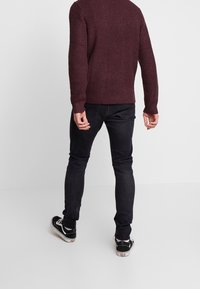 Maglione lavorato a maglia color porpora con superficie testurizzata, indossato sopra jeans neri aderenti e scarpe nere con suole bianche. Vista da dietro.
