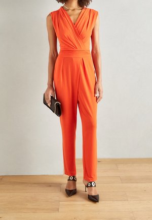 Combinaison orange vif avec un design portefeuille, sans manches et une taille cintrée. Associée à des talons pointus noirs ornés d'un accent circulaire.