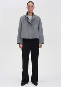 Graue Wolljacke mit hohem Kragen, doppeltem Knopfverschluss und weiten Ärmeln, kombiniert mit schwarzen geraden Hosen.