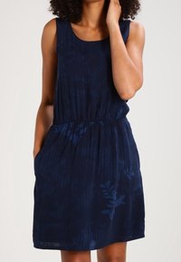 Woman wearing a sleeveless navy blue dress with subtle leaf patterns, hands in pockets, with curly hair partially visible.