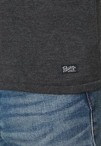 Dunkelgrau gestricktes Shirt mit strukturierter Oberfläche, versehen mit einem kleinen blauen Etikett am Saum. Trägt man zu hellblauen Jeans.