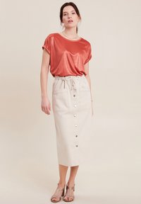 Roestrood metallic top met opgerolde mouwen, gecombineerd met een crèmekleurige midi-rok met knopen aan de voorkant en twee opgestikte zakken. Beige blokhak sandalen.