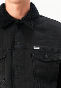 Crna traper jakna s teksturiranim crnim Sherpa ovratnikom, dugmadi na kopčanje, dva džepa na prsima i malim oznakom "Wrangler" sprijeda.