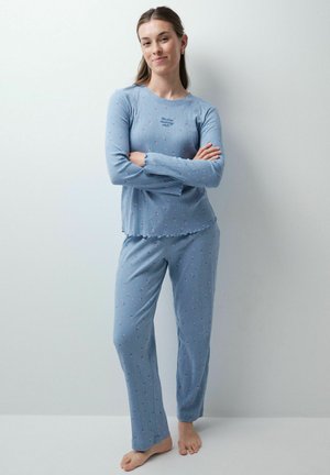 Jeune femme debout pieds nus, bras croisés, portant un haut bleu à manches longues et un pantalon avec de petits motifs floraux roses.