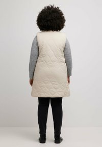 Beige gesteppte Weste mit hohem Kragen, seitlichen Taschen und mittellanger Länge bis zur Oberschenkelmitte, kombiniert mit einem grauen Langarmshirt und schwarzen Hosen.