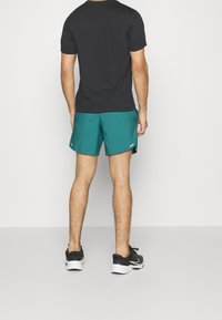 Svart t-shirt med kort ärm och teal atletiska shorts, med en jämn textur. Svarta sneakers med vita detaljer fulländar outfiten.