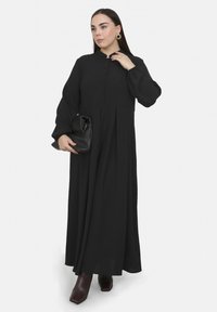 Schwarzes langärmliges Kleid mit Knopfleiste, plissiertem Rock und runder Halslinie. Modell hält eine schwarze Handtasche und trägt kastanienbraune Stiefeletten.
