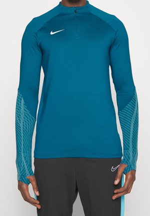 Mand iført en teal langærmet atletisk top med halv lynlås og mønstrede ærmer, kombineret med sorte bukser med hvidt Nike-logo.