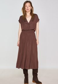 Abito maxi marrone a pieghe con scollatura a V e maniche corte; vita elastica; abbinato a stivali al ginocchio marrone scuro. Tessuto liscio e fluido.