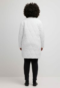 Bílý prošívaný dlouhý kabát s hladkým povrchem, s kulatým výstřihem a dlouhými rukávy. Kombinován s tmavými džíny a černými botami.