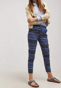 Blå mönstrade byxor med en blandning av geometriska och blommiga motiv, gjorda av lätt tyg, kombinerade med en ljusbeige jacka och sandaler.