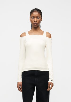 Mujer con cabello trenzado, vestida con blusa blanca de manga larga con hombros descubiertos y pantalones negros, de pie frente a un fondo claro y liso.