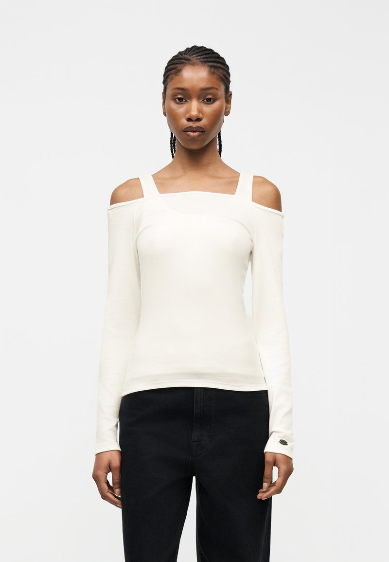 Γυναίκα με πλεγμένα μαλλιά, που φοράει λευκή μπλούζα με ανοιχτούς ώμους και μακρύ μανίκι και μαύρο παντελόνι, στέκεται μπροστά σε απλό φωτεινό φόντο.