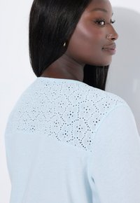 Světle modrá košile s květinovým krajkovým vzorem na zadním dílu a měkkou bavlněnou texturou. Kulatý výstřih a dlouhé rukávy.