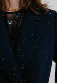 Femme portant un manteau bleu marine foncé avec de petits sequins dispersés sur un haut noir transparent orné de sequins floraux noirs et argentés.