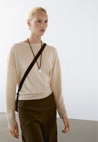 Maglione lavorato a maglia beige con vestibilità comoda, colletto rotondo e maniche lunghe, indossato con pantaloni a vita alta marrone scuro e una collana con pendente.