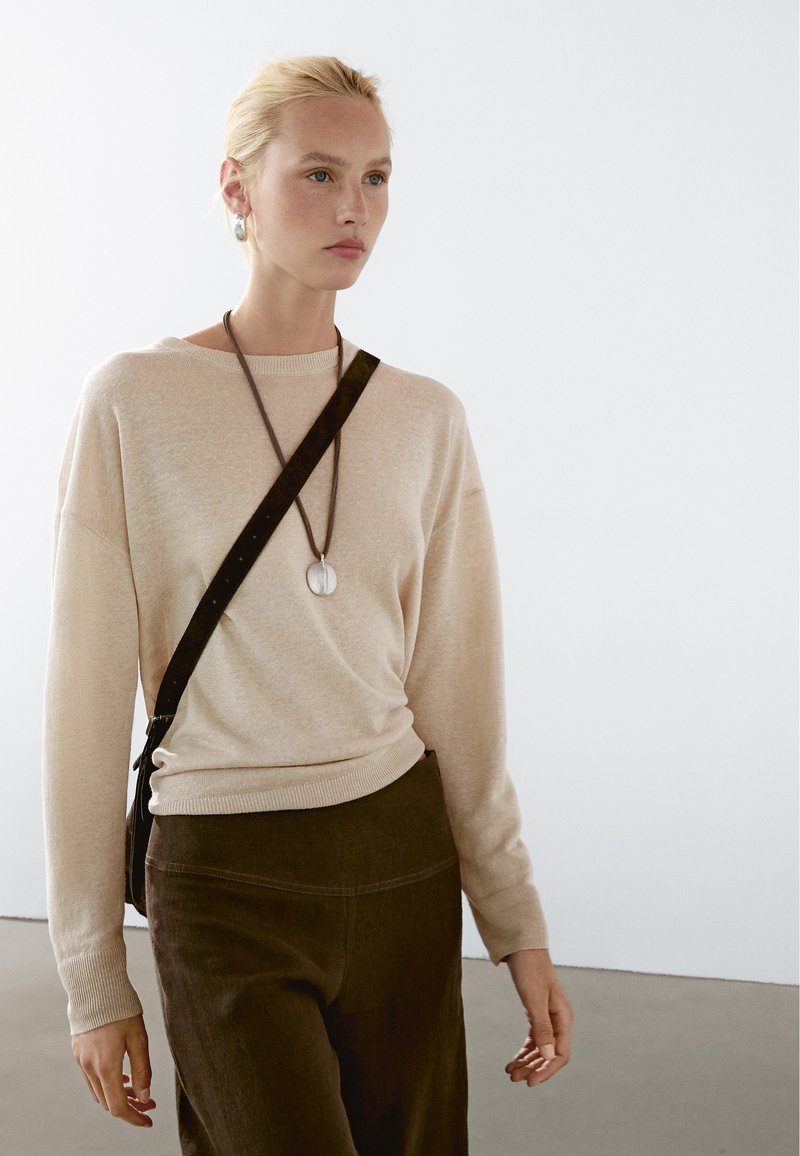 Maglione lavorato a maglia beige con vestibilità comoda, colletto rotondo e maniche lunghe, indossato con pantaloni a vita alta marrone scuro e una collana con pendente.