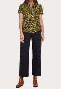 Chemise à manches courtes jaune avec imprime léopard noir, associée à un pantalon noir à jambes larges et des bottines à lacets en simili-crocodile marron.