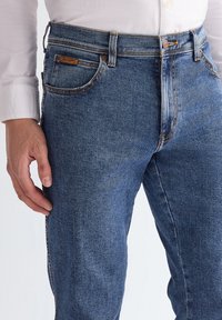 Férfi kék farmernadrágot visel, amelyen öt zseb található, és fehér hosszú ujjú inget, keze a combja mellett pihen.