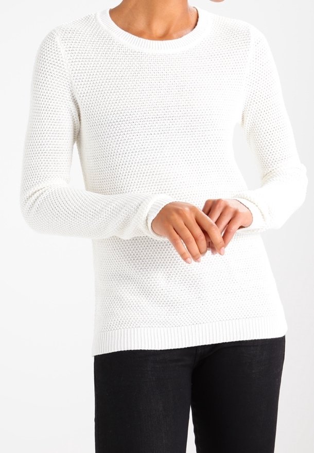 Personne portant un pull en maille blanc texturé à manches longues et un pantalon noir, les mains jointes de manière détendue devant.
