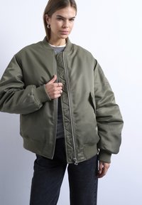 Jeune femme portant une veste bomber oversize vert olive sur des hauts superposés, debout devant un fond clair uni.