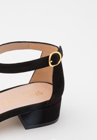Sandale basse en daim noir avec bride de cheville à boucle dorée et semelle intérieure beige avec lettrage doré sur fond blanc.
