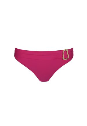 Vrouw met een lichte huid en haar in een staart, draagt een magenta bikini met gouden gespdetails, staand tegen een eenvoudige lichte achtergrond.