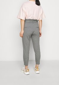 Žena stojící zády v světle růžovém tričku s krátkým rukávem, vysokých šedých kalhotách a bílých teniskách se béžovými akcenty.