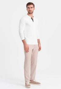 Weißes Langarm-Henley-Shirt mit schwarzen Knopfakzenten, kombiniert mit hellbeigen Hosen und grauen Sneakers. Glattes Material und taillierte Passform.