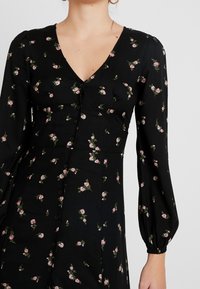 Sort kjole med lange puffærmer, V-hals og knapper foran, med et blomstermønster af små lyserøde roser og grønne blade.