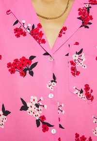 Rosa blommig skjorta med knappar som har röda och vita blommor, korta ärmar, en bröstficka och krage med tänder i en mjuk textur.