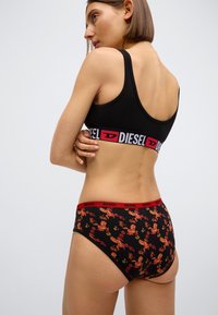 Černá sportovní podprsenka s výrazným červeným páskem s logem Diesel, doplněná o černé kalhotky s vysokým pasem s výrazným květinovým vzorem v červené a zlaté barvě.