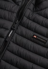 Chaqueta acolchada negra con textura de acolchado, con cremallera frontal y un parche del logotipo de Superdry en rojo y blanco en el lado izquierdo.