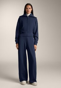 Nainen, jolla on yllään tummansininen vetoketjullinen collegetakki ja leveälahkeiset housut sekä valkoiset kengät, seisomassa yksinkertaisen beigen taustan edessä.