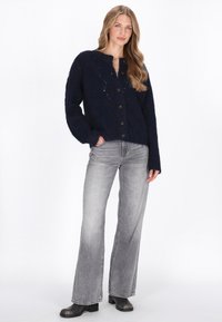 Tengerészkék kötött kardigán kerek nyakkivágással, nagy gombokkal és laza ujjakkal, szürke trapézfazonú farmernadrággal és fekete bokacsizmával párosítva.