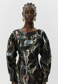 Moteris su pintomis plaukų uodegomis, vilkinti juodą suknelę su mėlynomis, rožinėmis ir auksinėmis abstrakčiomis raštomis, bei didelėmis auksinėmis ir perlinėmis pakabinamomis auskarėmis.