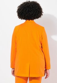 Oranssi räätälöity bleiseri, jossa on jämäkkä malli, lovet lapel-it ja halkio takana. Yksi nappisulku ja näkyvää yksityiskohtaista ompelua.