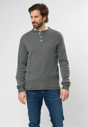 Siva dugmadna Henley majica s dugim rukavima i teksturiranim dizajnom, tri gumba na ovratniku i mali logo na prsima. Nosi se s plavim trapericama.